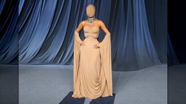 Kim Kardashian in a tan dress and face covering with her hands on her hips.