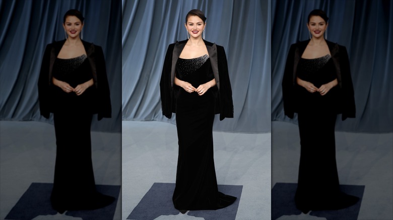 Selena Gomez in a black dress with a black blazer draped over her shoulders in front of a blue backdrop