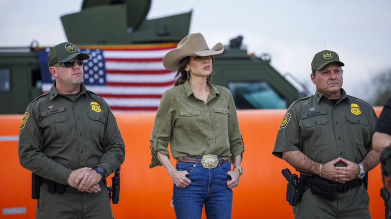 Kristi Noem wearing a tan cowboy hat, green shirt, and blue jeans and posing between two border patrol agents.