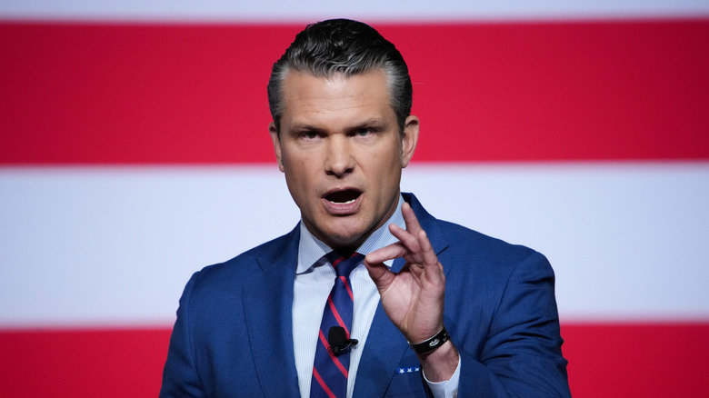 Pete Hegseth at an event wearing a a dark blue suit and gesturing with hands while speaking
