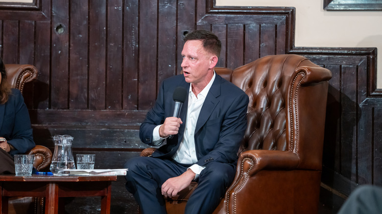 Peter Thiel sitting in a leather chair and speaking into a microphone at an event in Cambridgeshire, United Kingdom