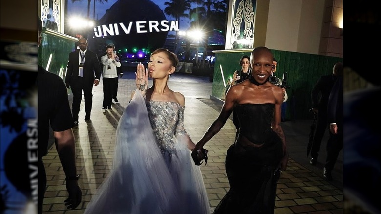Ariana Grande blows a kiss in a blue gown while holding hands with Cynthia Erivo as they walk down a yellow brick carpet.