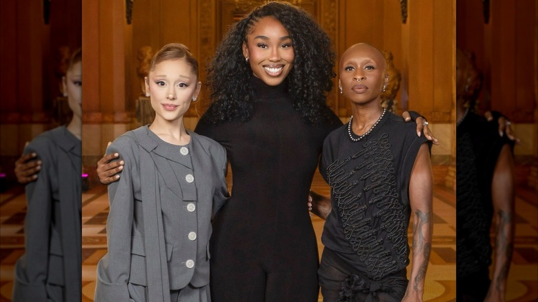 Ariana Grande, Sally, and Cynthia Erivo in a gold hall.