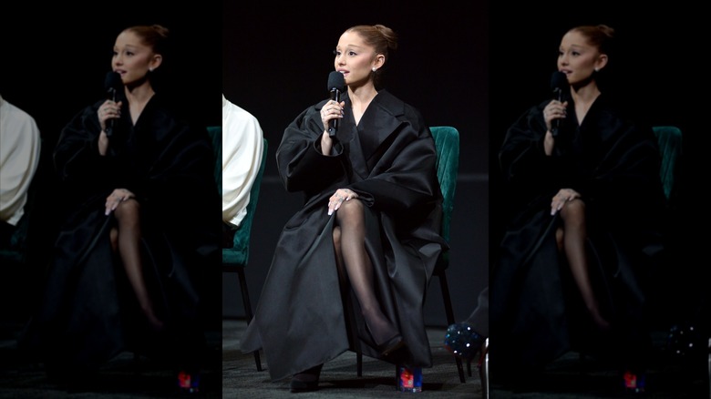 Ariana Grande in all black, sitting in a green chair, holding a microphone to her mouth.