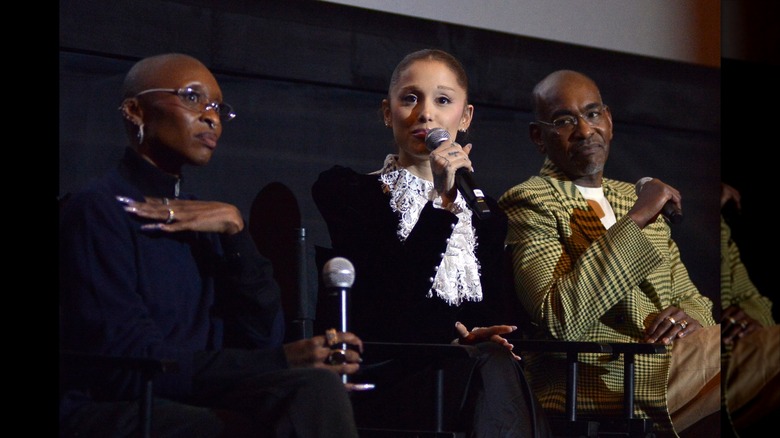 Ariana Grande speaking into a microphone beside Cynthia Erivo.