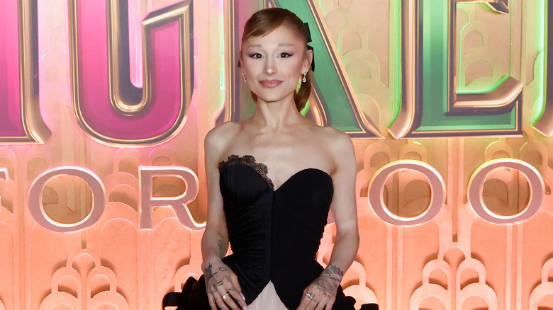 Ariana Grande in a black and pink dress in front of a wicked sign.