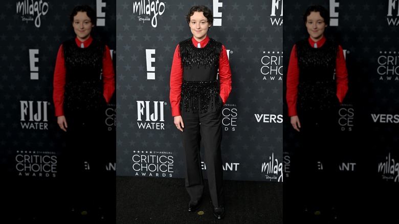 Bella Ramsey posing in a red shirt and black jumpsuit with beaded fringe.