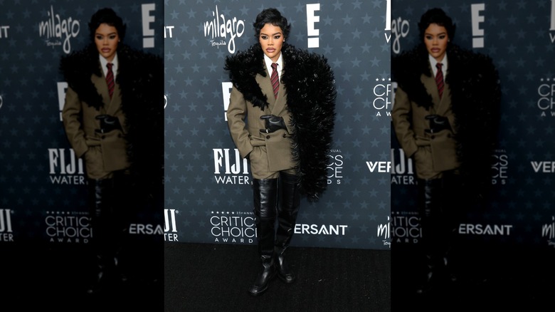 Teyana Taylor posing in a tan double-breasted coat with a large feather detail and black-thigh boots.