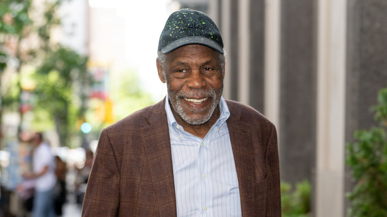 Danny Glover smiling in a brown plaid jacket, light blue button-up, and black hat with green speckles on the streets of Philadelphia in 2025