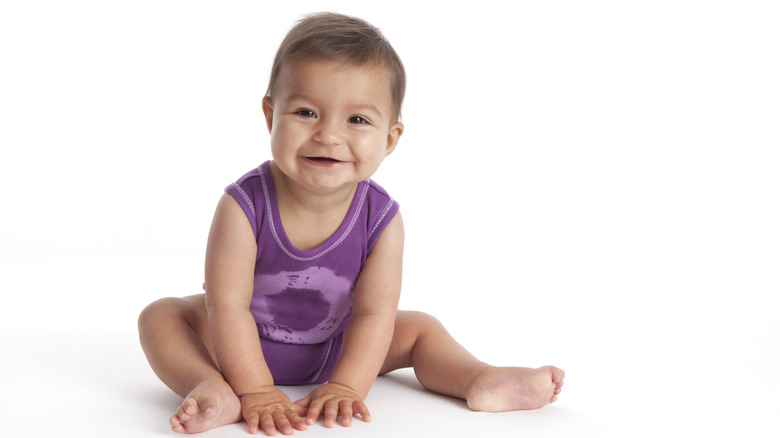 A baby girl smiling and sitting with her hands on the ground between her legs