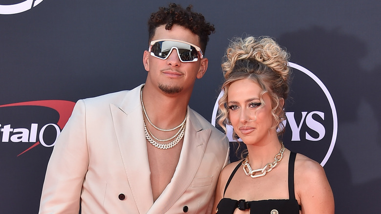 Patrick Mahomes wearing sunglasses, gold chains, and a beige suit jacket and Brittany Mahomes wearing a black dress posing together on the ESPY red carpet in 2023.