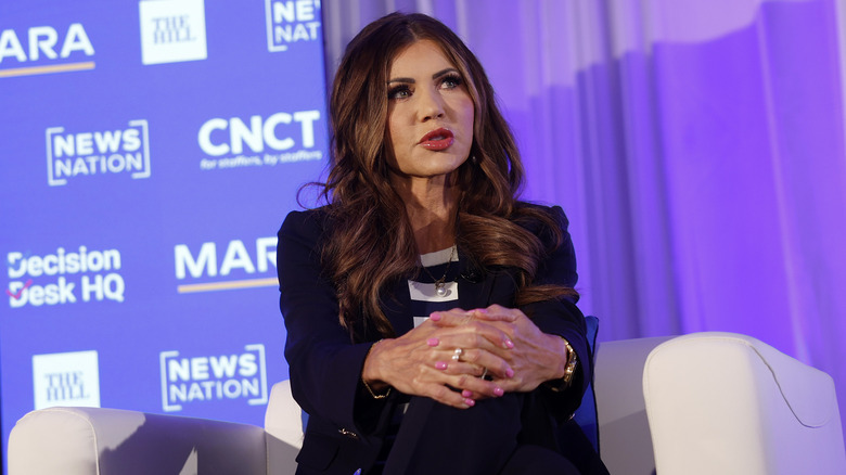 Kristi Noem speaking while clasping her hands together in a black blazer and stripy monochrome top