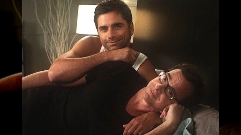John Stamos and Bob Saget lay in a bed together, cuddling and smiling.
