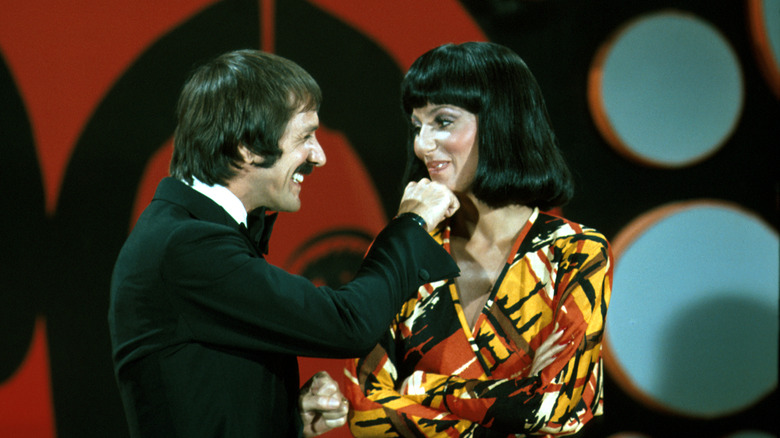 Sonny Bono smiling and holding his fist at Cher's chin