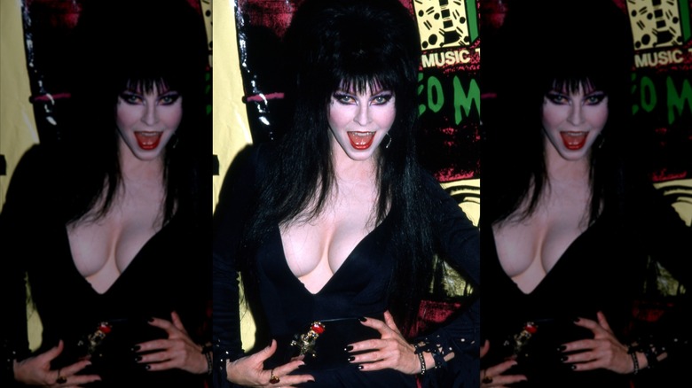 Elvira wearing a black dress and dagger belt while posing with her hands on her waist at the 1988 MTV VMAs.