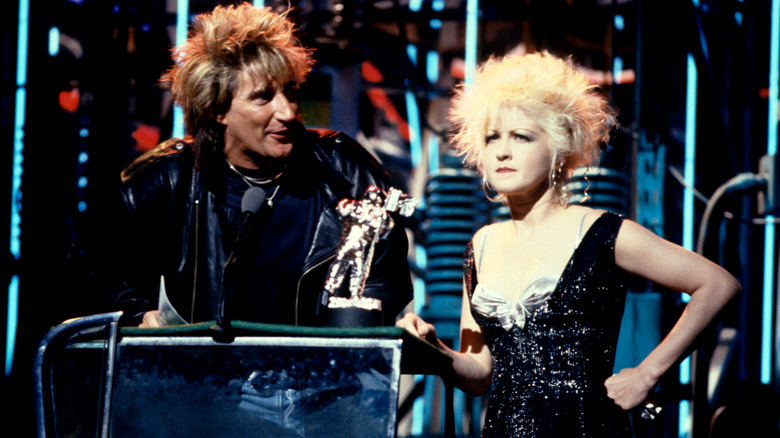 Rod Stewart speaking on stage at the 1988 MTV Video Music Awards while Cyndi Lauper frowns beside him.