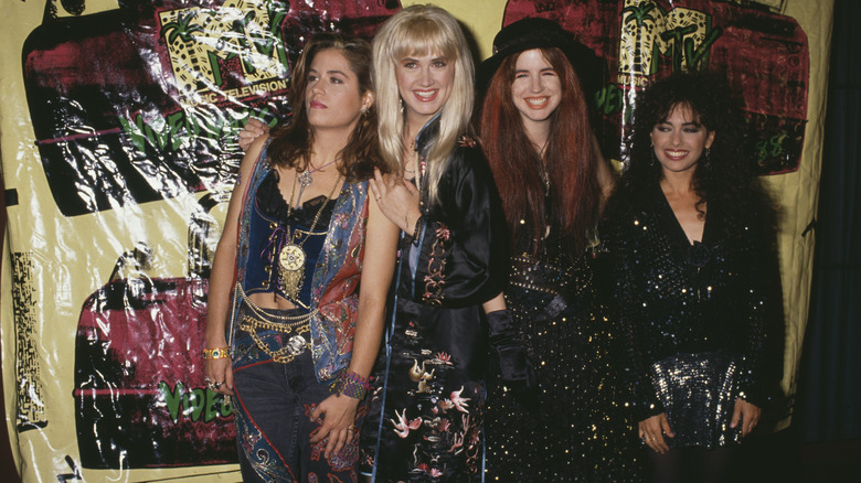 Vicki Peterson wearing a patterned vest, denim bustier, embroidered jeans, and gold accessories while posing with her bandmates at the 1988 VMAs.