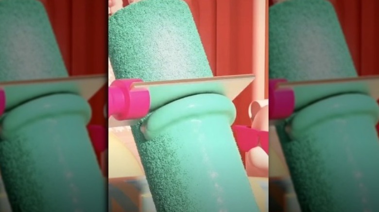 Paint roller filled with green paint with a scraper running over it to remove the paint.