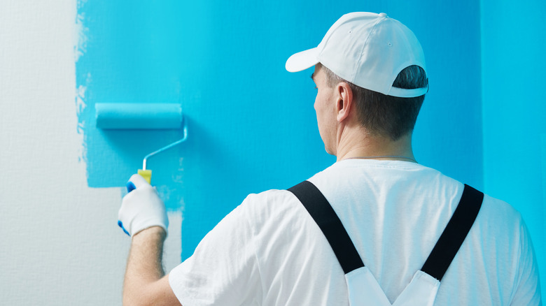 Man wearing white facing a wall and painting it blue with a paint roller.