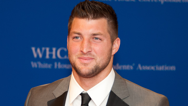 Tim Tebow smiles while wearing a tan suit and tie.