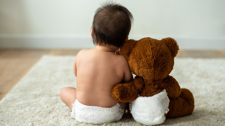 Baby leaning against a teddy bear from the back