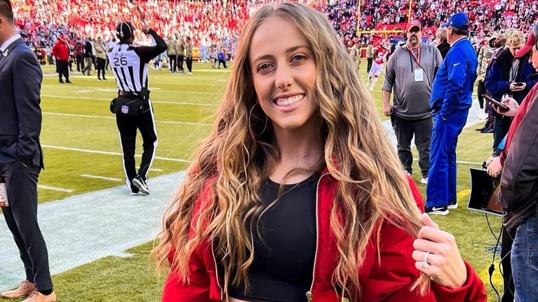 Brittany Mahomes on football field sidelines smiling at camera, with dark blonde hair and red jacket