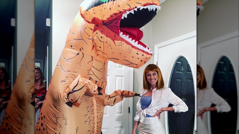 Brittany Mahomes dressed as Jurassic Park figure in red wig, standing next to giant inflatable dinosaur