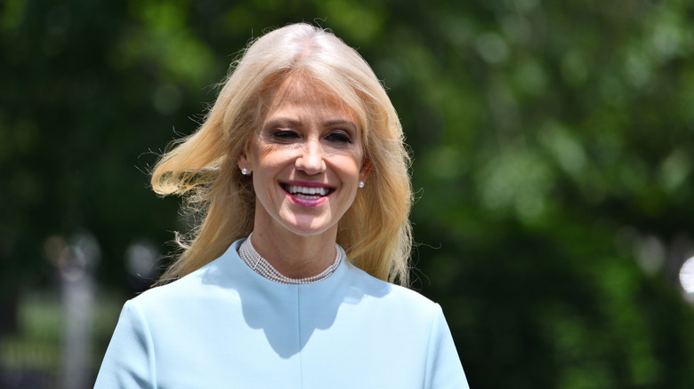 Kellyanne Conway in light blue outfit smiling outdoors, with greenery in the background