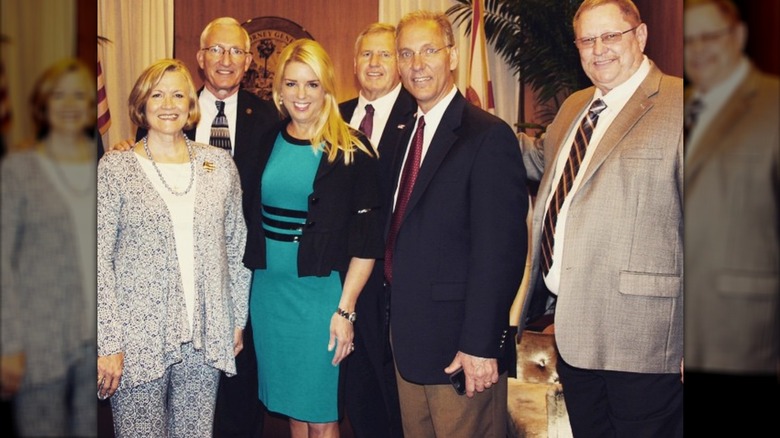 Pam Bondi wears a teal sheath dress with black stripes and a cropped black jacket while posing with a group of people in the Florida attorney general's office.