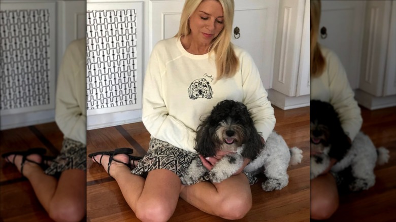 Pam sits on the floor with a dog while wearing a black and white patterned skirt and a sweatshirt with a drawing of the dogs face on it.