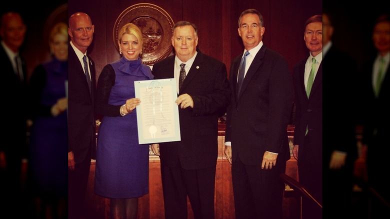 Pam Bondi wears a purple dress with scarf detail over a black long-sleeved shirt and pantyhose in a group photo.