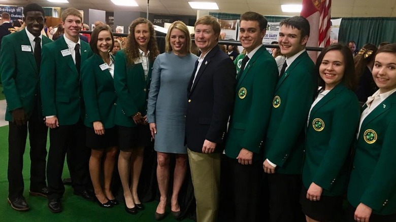 Pam Bondi wears a wrinkled long-sleeved blue dress and poses with 4-H members wearing green blazers.