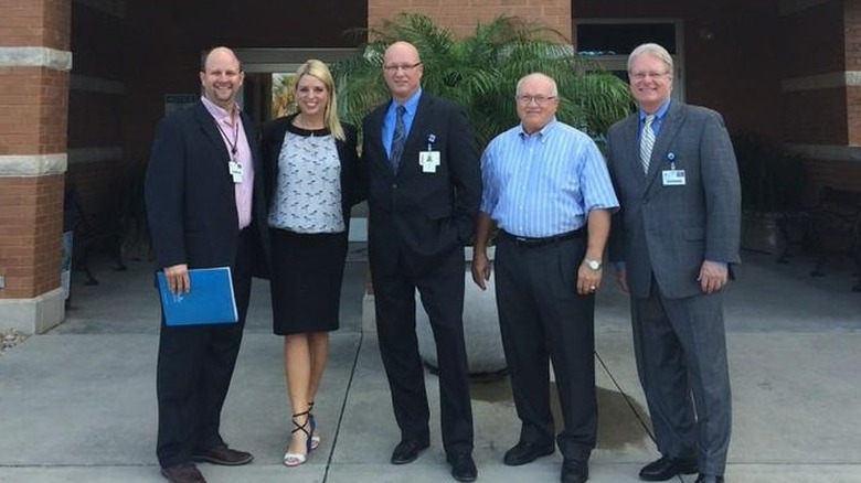 Pam Bondi wears strappy high heels, a black skirt, black blazer, and white patterned shirt while posing with a group of men outside a medical facility.