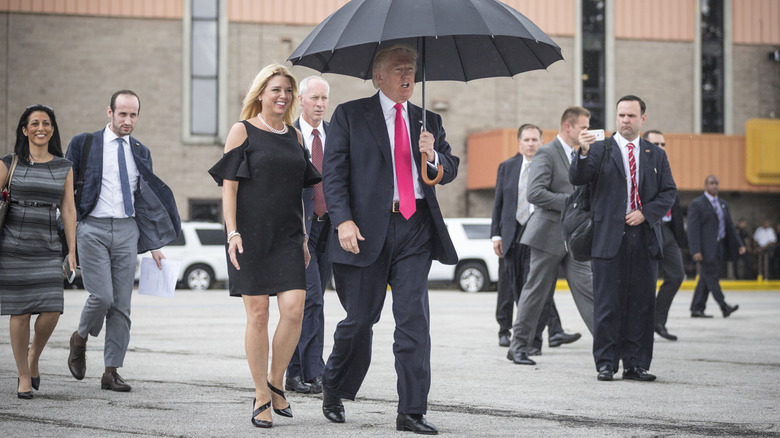 Pam Bondi wears a black minidress with ruffled sleeves and walks beside Donald Trump, who is holding an umbrella.