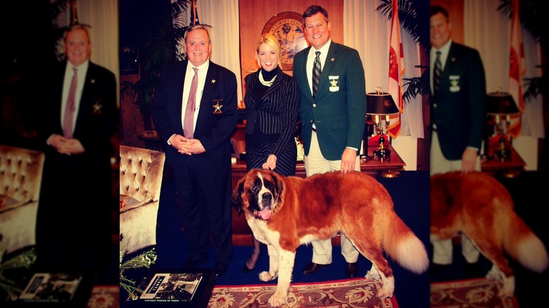 Pam Bondi wears a black pinstripe suit dress and strings of pearls while posing with two men and a dog in the Florida attorney general's office.
