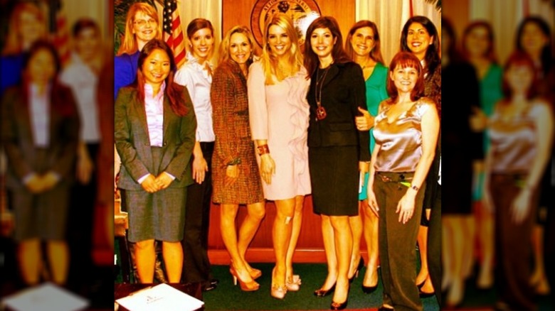 Pam Bondi wears a pink dress with a large ruffle down the front while posing with a group of women inside the Florida attorney general's office.