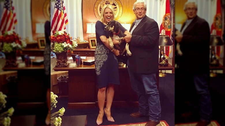 Pam Bondi wears a dark blue sweater dress with white floral designs on the hips and holds a dog in the Florida attorney general's office.