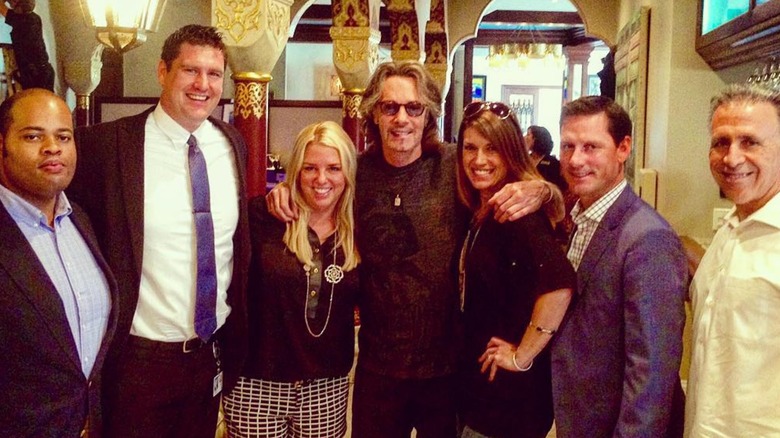 Pam Bondi wears black shorts with a white grid print and a black polo shirt while posing with Rick Springfield and others.