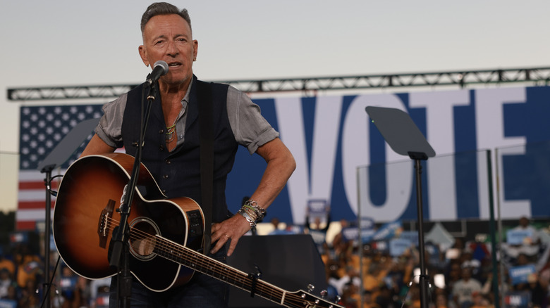 Bruce Springsteen stands on stage with his guitar with a "Vote" banner in the background during a rally for Kamala Harris.