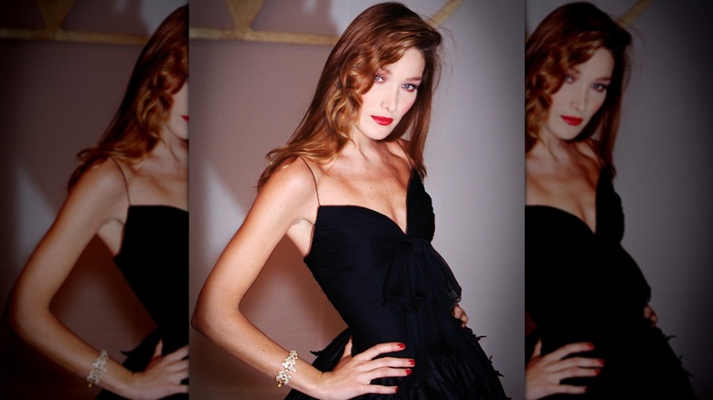 Carli Bruni poses with her hands on her hips backstage at Paris Fashion Week in 1994 in a black dress, bright red lipstick, and deep side part.