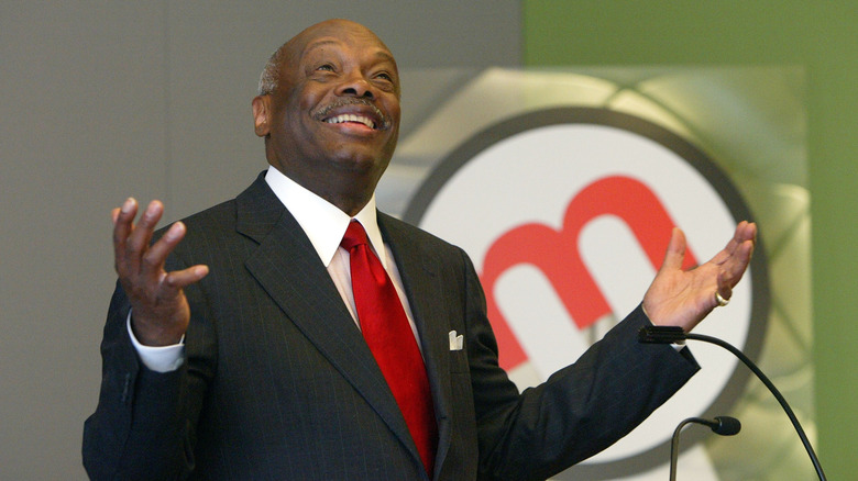 Willie Brown wears a red tie and looks up with his arms outstretched.