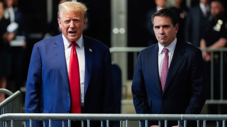 Todd Blanche looking serious as he stands alongside a talking Donald Trump