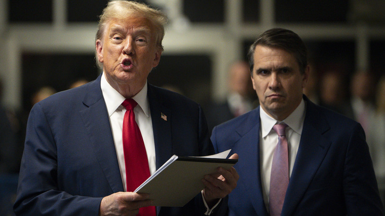 Donald Trump speaking while holding a pile of papers as Todd Blanche looks on with a stormy expression