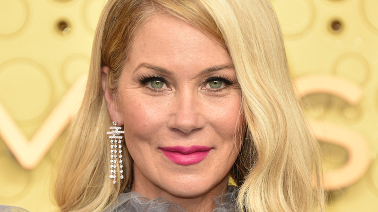 Christina Applegate smiling while posing at an event in a grey dress with dangly earrings