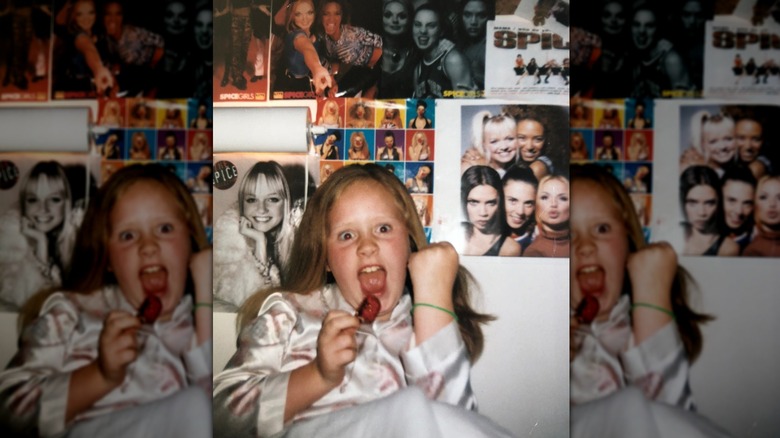 Adele as a kid in her pajamas singing into a red pacifier pop in front of Spice Girls posters on the wall behind her