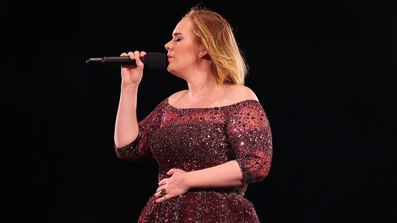 Adele in red, sparkly dress on stage singing into microphone with her eyes closed