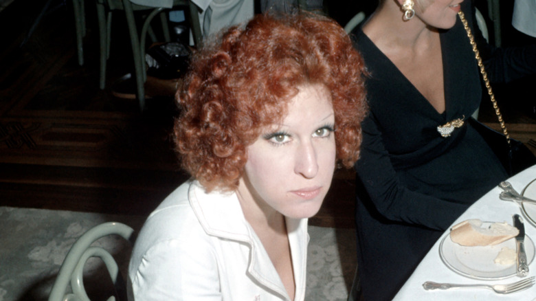 Young Bette Midler with red curls, sitting at a table, looking up