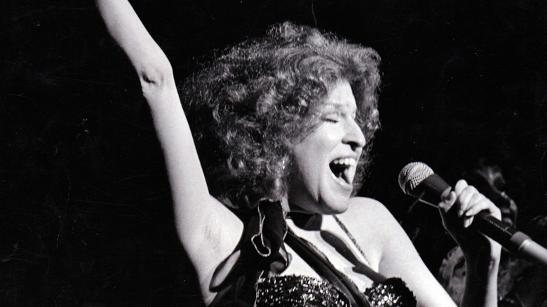 Bette Midler singing into a microphone with her arm up in the air, in black and white