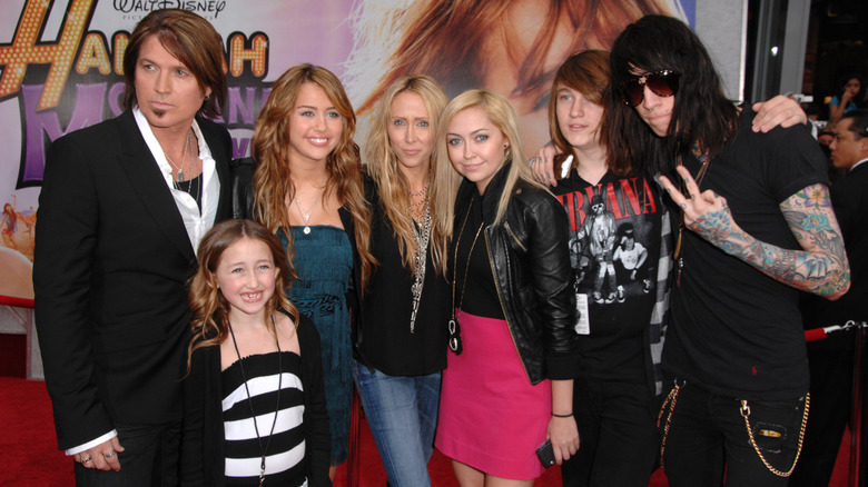 Billy Ray Cyrus in black suit with wife Tish in black shirt and jeans and his five children in front of Hannah Montana poster