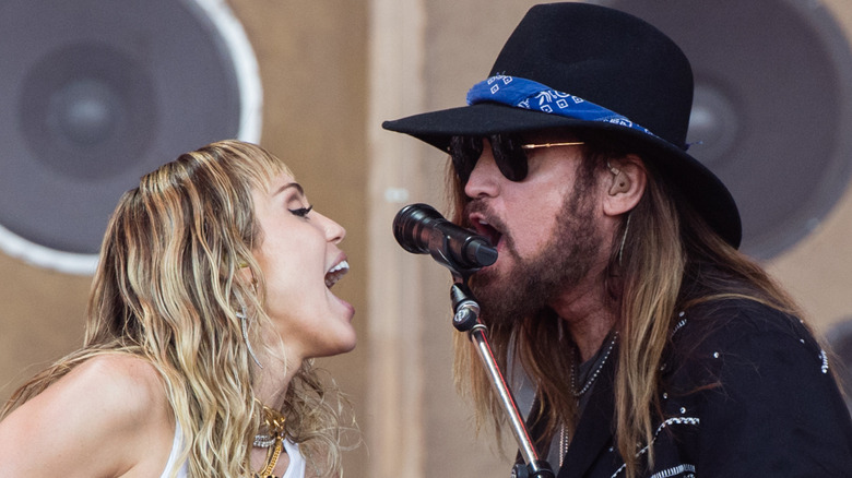Billy Ray Cyrus in black hat and shirt and Miley Cyrus in white crop top and black pants performing together